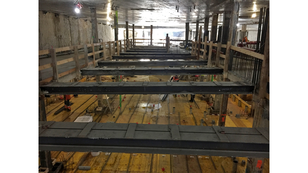 Temporary steel beams secure structural work in the excavation pit in the area of what will become a multistory building-services unit. Premade sheet pile walls can be seen framing the pit on the side.